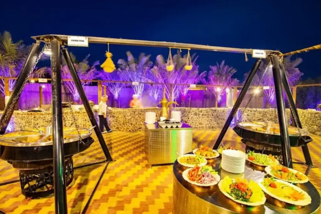 BBQ buffet set up at a desert camp at night with tents lit in purple